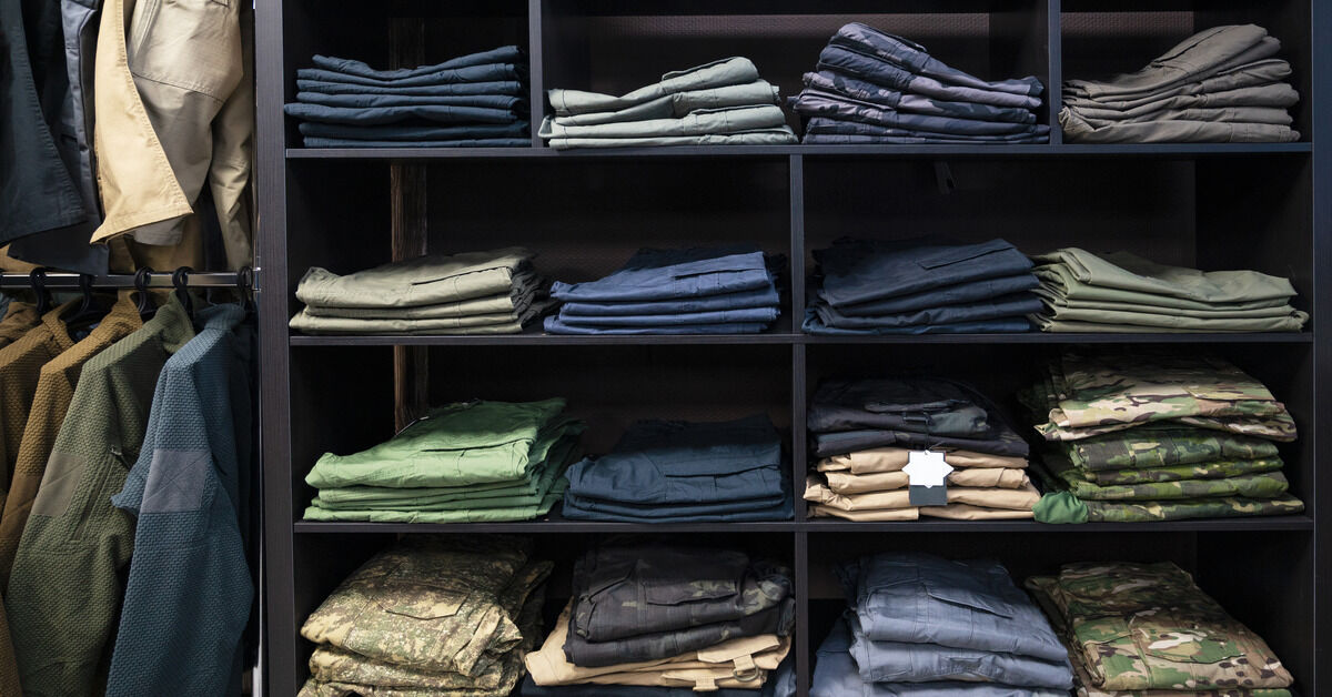 A black shelf with blue, green, beige, and camouflage pants, each neatly folded, next to a rack of military shirts on hangers.